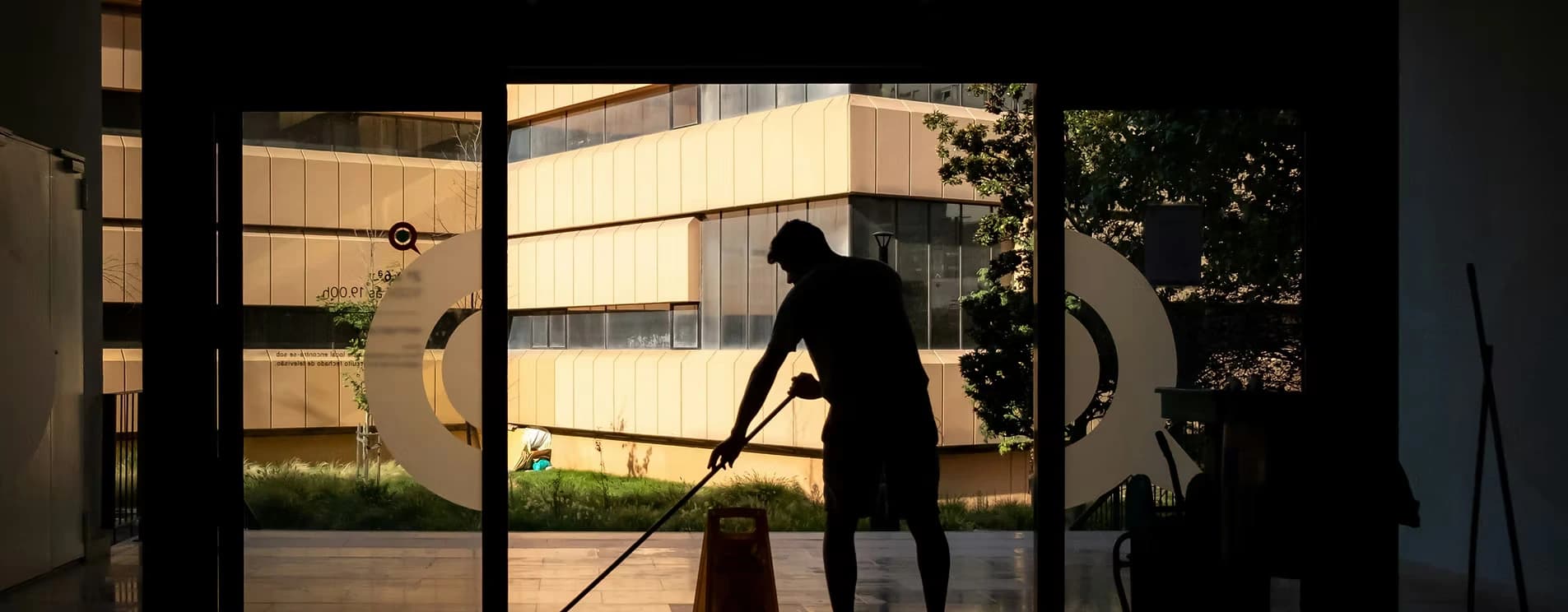 Ein Putzmann als Silhouette, der bei Sonnenuntergang oder starkem Gegenlicht den Boden in einem gläsernen Eingangsbereich wischt. Im Hintergrund ist ein modernes, hellbraunes Gebäude zu sehen.